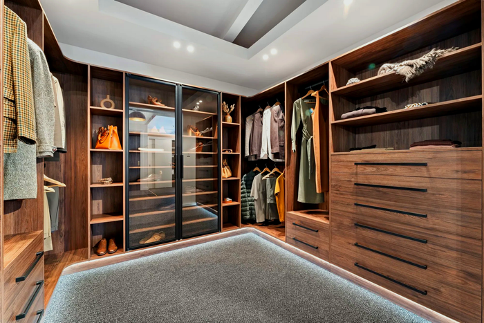 A luxury walk-in closet with wooden shelving, glass-door display, and a grey rug with built-in lighting. Clothes, shoes, and accessories are organised within.