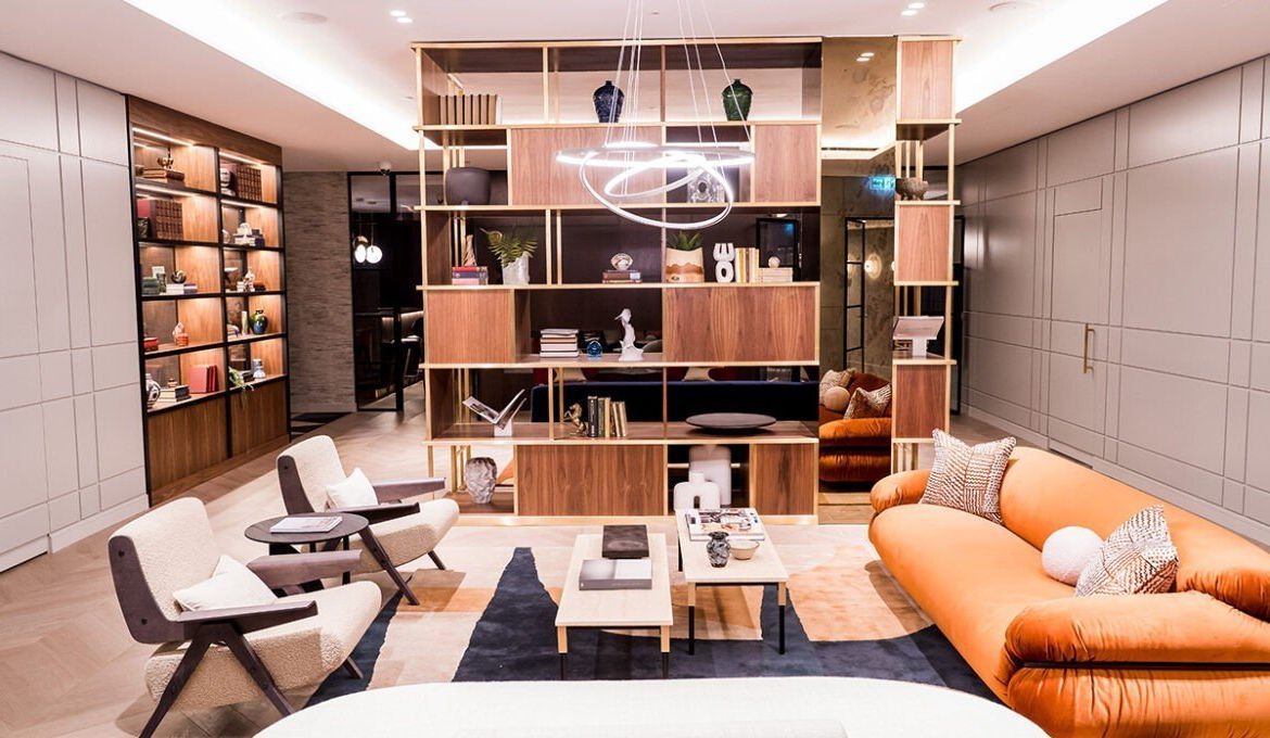 Modern office lounge with orange sofa, white chairs, coffee table, and wall shelving.