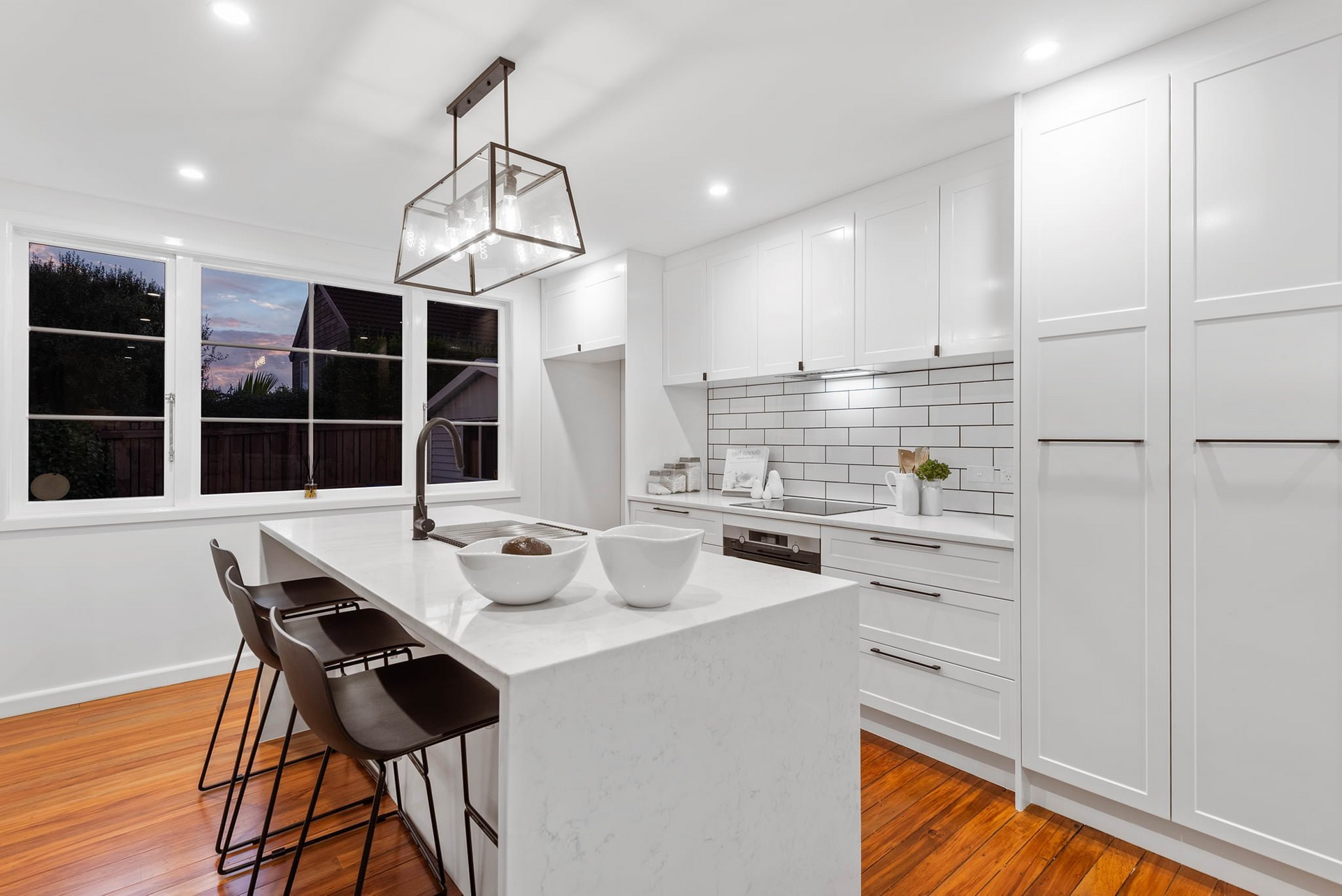 Modern white kitchen with island, dark stools, and hardwood floors, illuminated by a geometric chandelier.