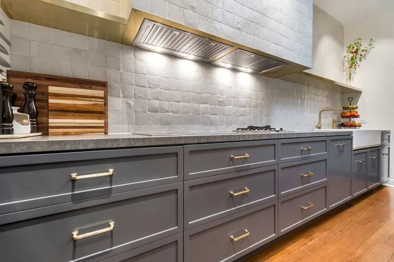 Gray kitchen cabinets with gold handles under a white tiled backsplash, stainless steel hood, and wooden countertop.