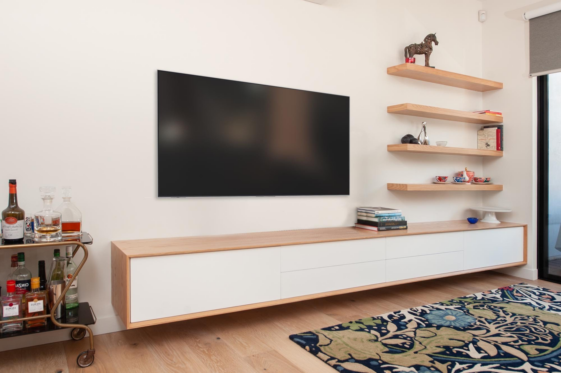A modern living room with a wall-mounted TV and floating shelves. A white and wood cabinet sits below the TV. A bar cart is on the left, and a rug is on the floor.