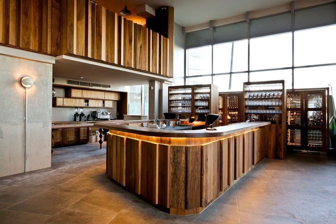 Modern restaurant interior with a wooden bar counter, shelves, and large windows.
