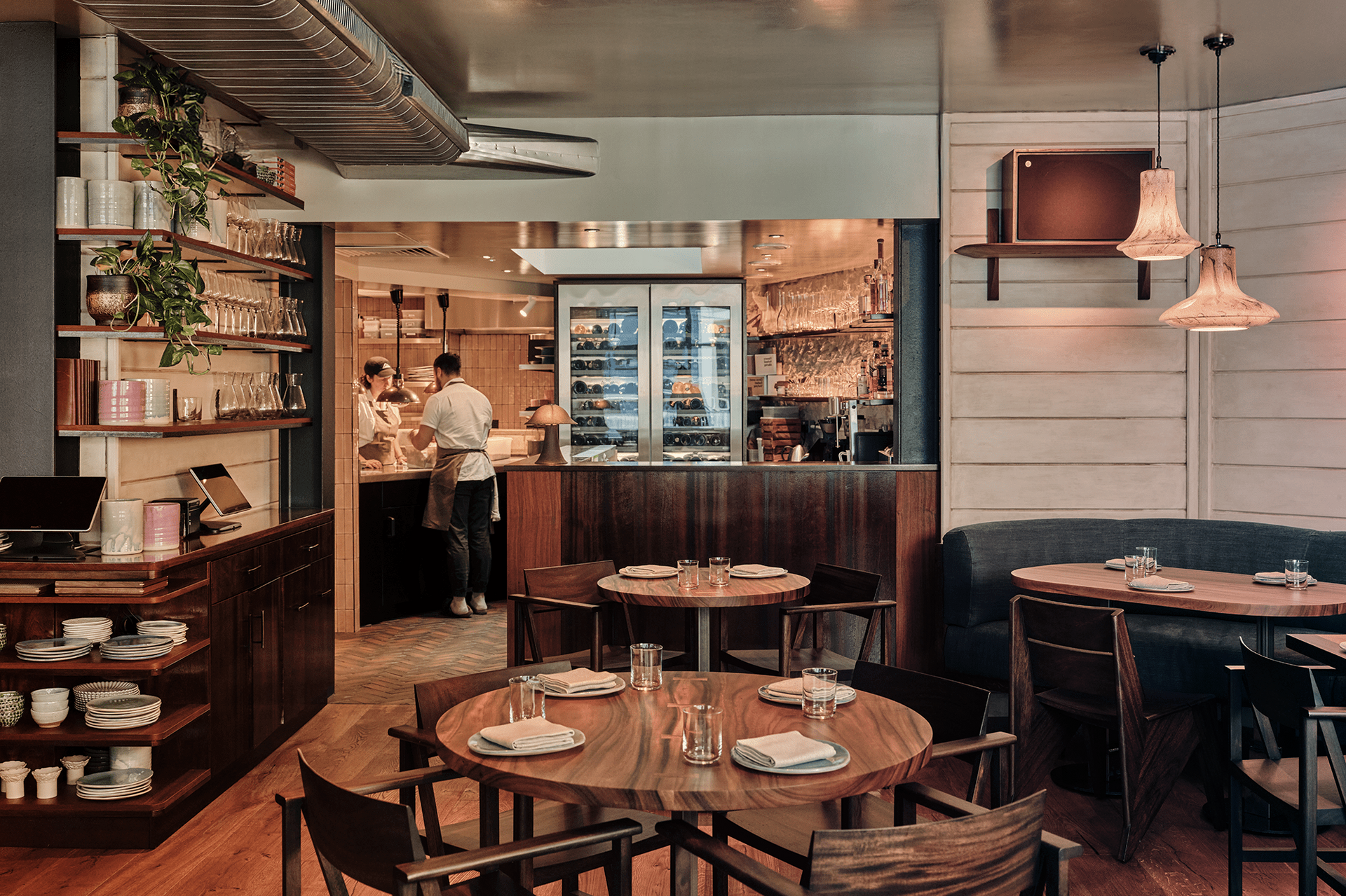 Cozy restaurant interior with wooden tables, shelves, hanging lights, and a chef working in the back kitchen.