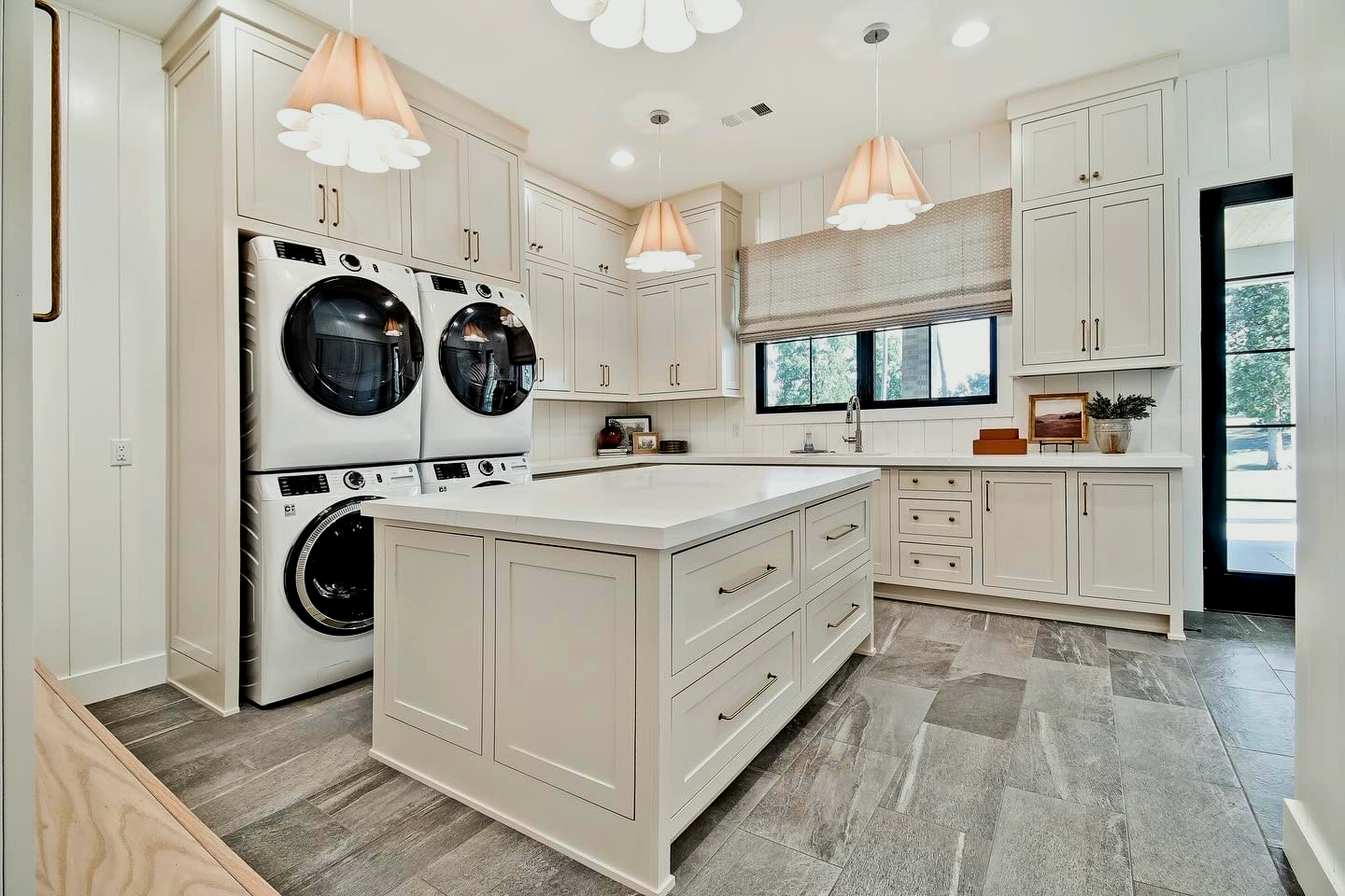 A white laundry room with cabinets, island, washer/dryer, and gray stone floor.  Three pendant lights and a window.