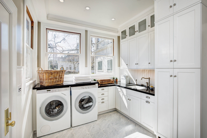 White laundry room with washer and dryer, cabinets, and a sink by two windows.