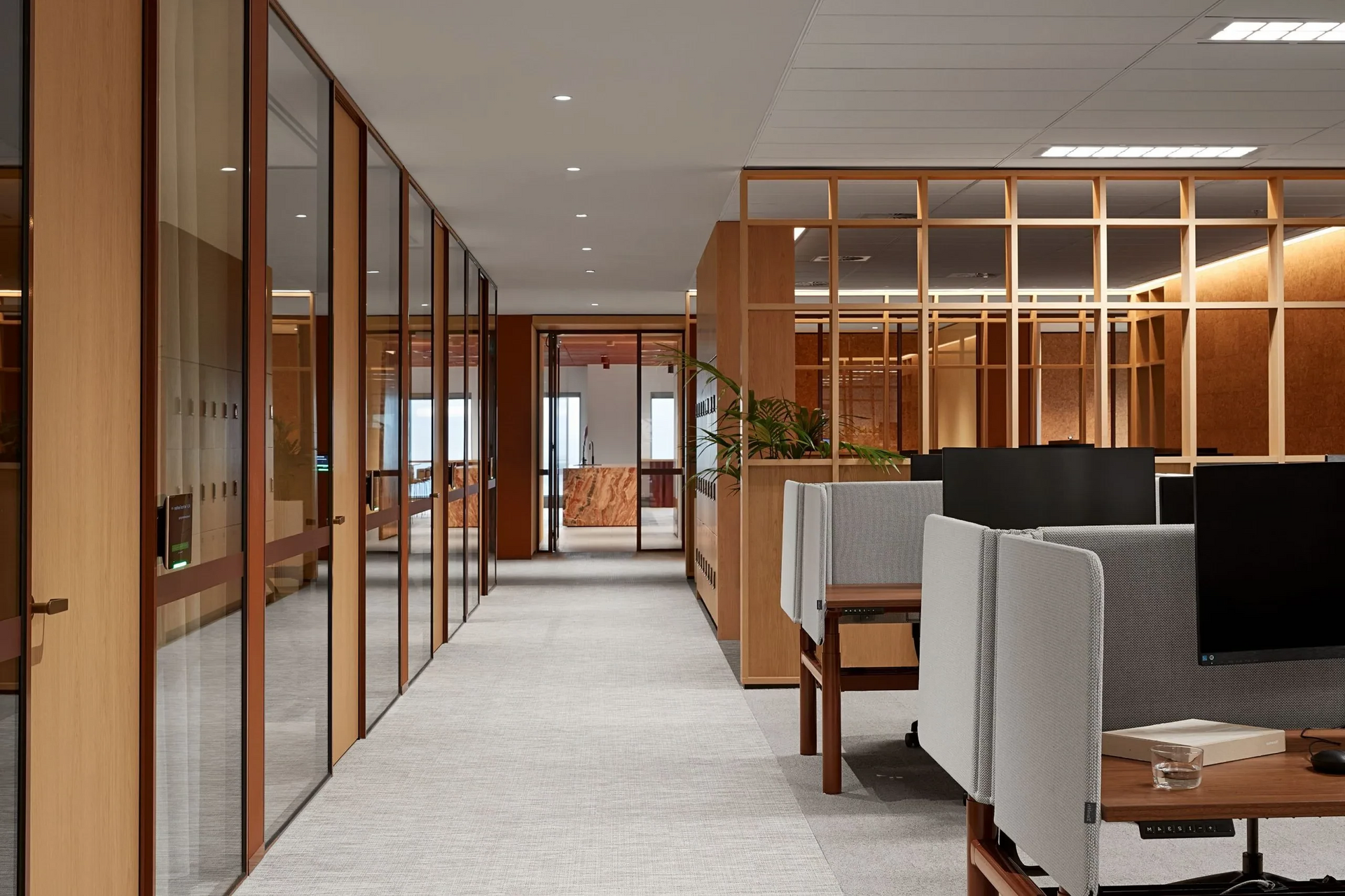 Modern office hallway with glass walls, carpeted floor, and seating area with wood accents.
