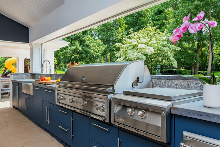 Outdoor kitchen with stainless steel grill, griddle, and sink, against a blue cabinet, and green foliage in the background.