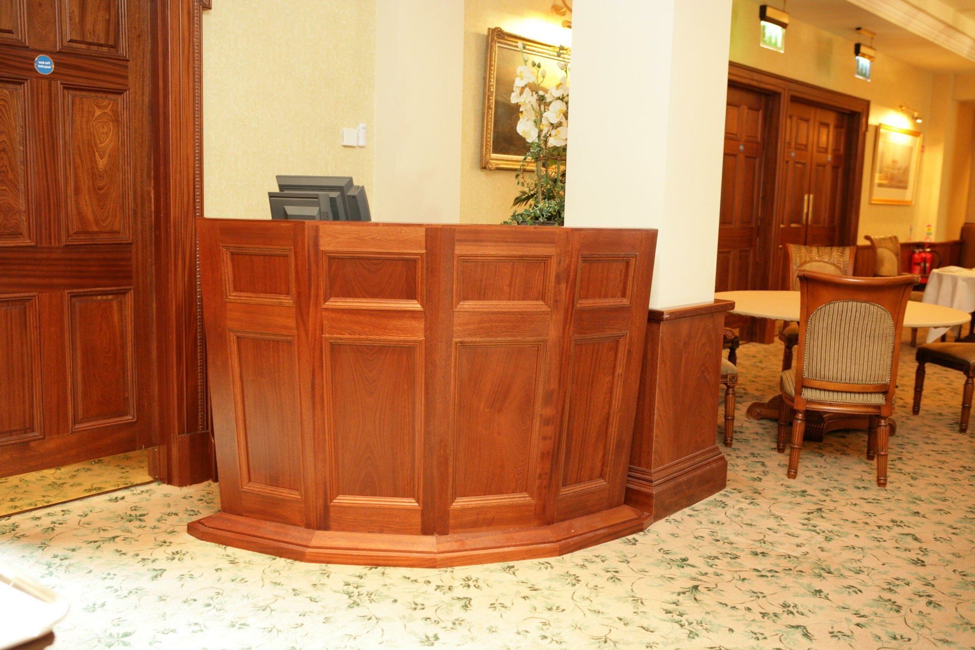 Wooden hotel reception desk in a warm-lit lobby with patterned carpet and chairs nearby.