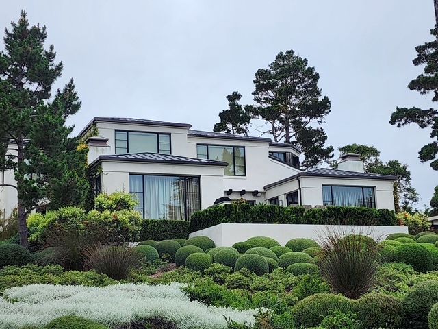 White modern house with multiple levels, surrounded by manicured green shrubs and trees, under an overcast sky.