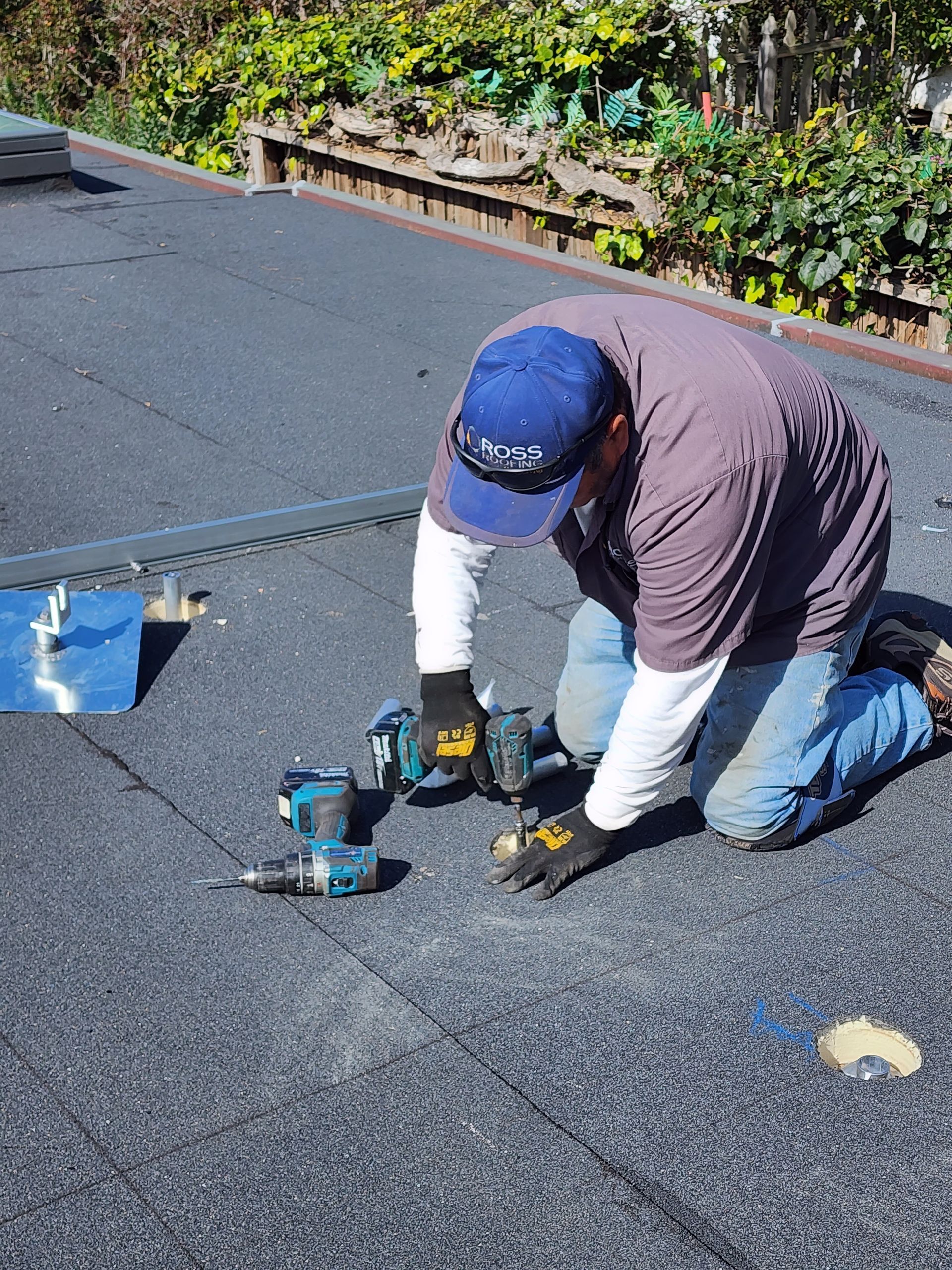 Person kneels on a black rooftop, using power tools. Blue hat, gray shirt, and gloves are visible. Setting is outdoors.