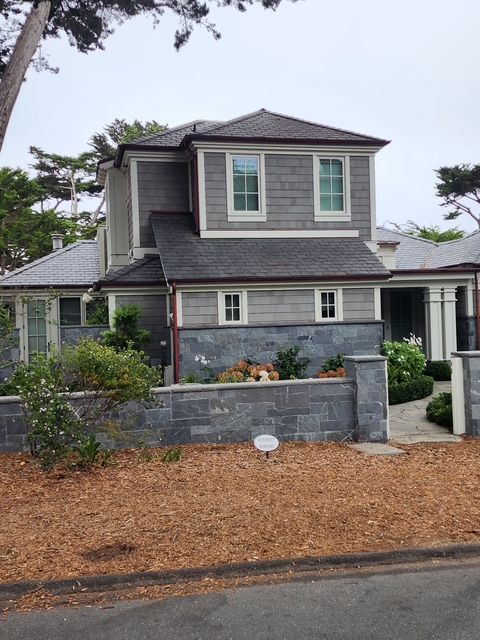 Two-story gray house with wood shingle siding and dark roof, behind a stone wall with mulch.