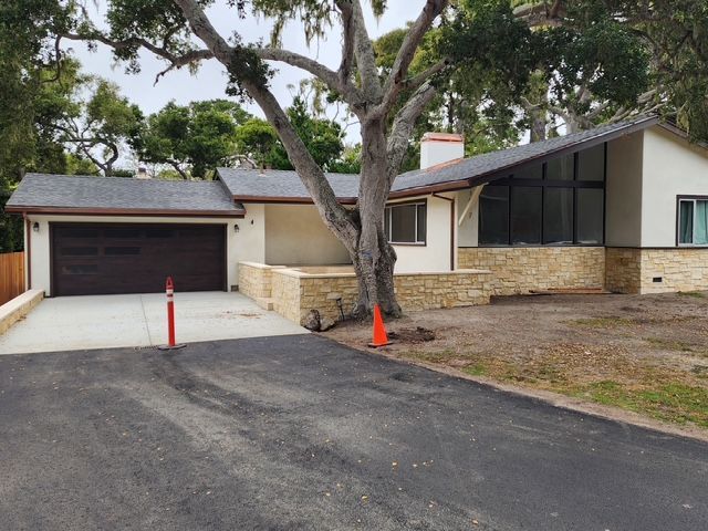 A one-story house with a tree growing in front; a black driveway and brown garage door are visible.