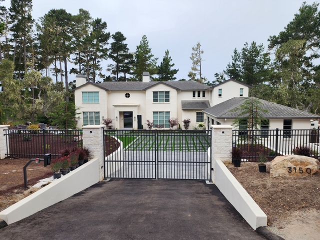White house with black gate and fence, surrounded by trees. Cloudy sky.
