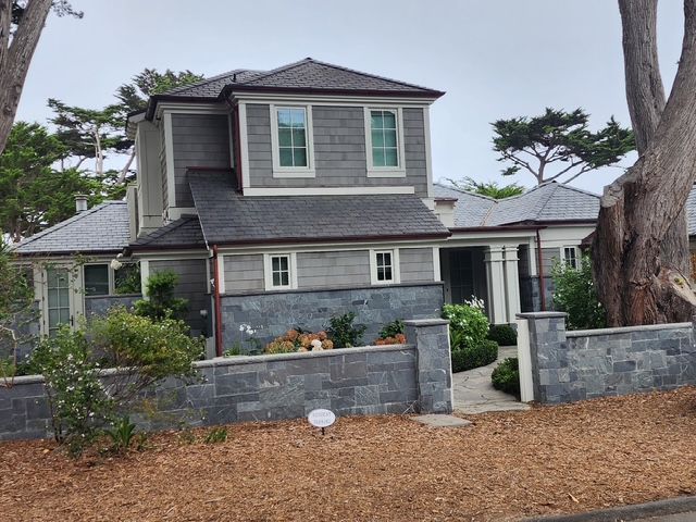 Gray two-story house with stone and shingle exterior. Features a small front yard, stone wall, and overcast sky.