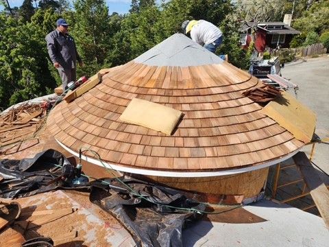 Workers installing wooden shingles on a round roof. One stands above, another to the left.