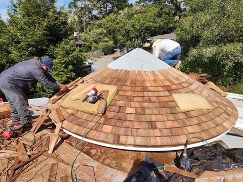 Two workers installing cedar shingles on a domed roof in a wooded area.