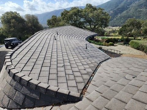 Curved roof with gray shingles. A black vehicle parked next to the roof, mountains in the background.