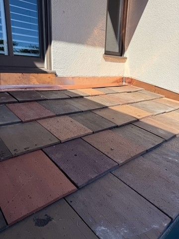 Close-up of a tiled roof in various brown and reddish tones, adjacent to a beige wall and window.
