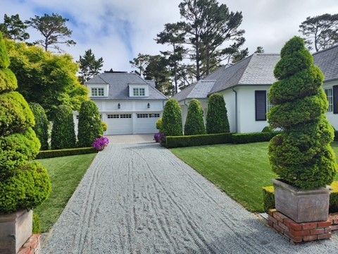 Driveway leading to a white house with manicured hedges and topiary. Overcast sky.