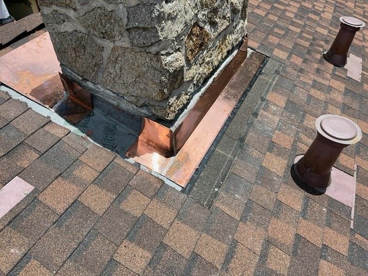 Close-up of a chimney on a shingled roof with copper flashing for water protection and two vent pipes.