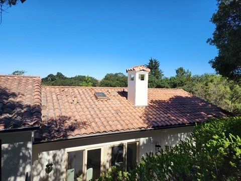 Red-tiled roof of a house with a white chimney against a blue sky, surrounded by green trees.