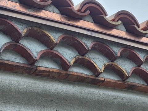 Close-up of a tiled roof edge with red-brown clay tiles in a wave pattern against a light gray wall.