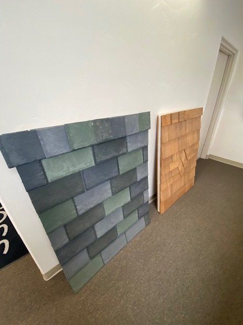 Two roofing shingle samples: grey/green brick pattern and brown wood-like texture, on a wall and carpet.