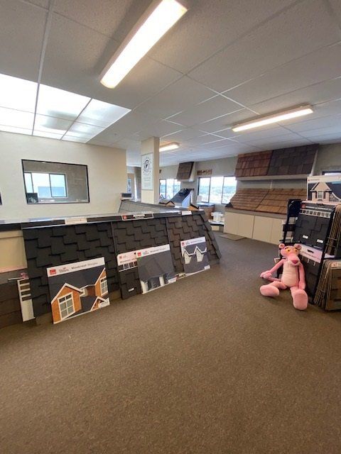 Interior of a roofing supply showroom. Samples of shingles on display, pink panther plush toy, and fluorescent lighting.