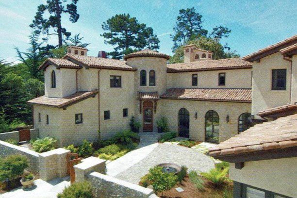 Beige stucco house with a red tile roof, and a circular tower, set among trees and greenery.