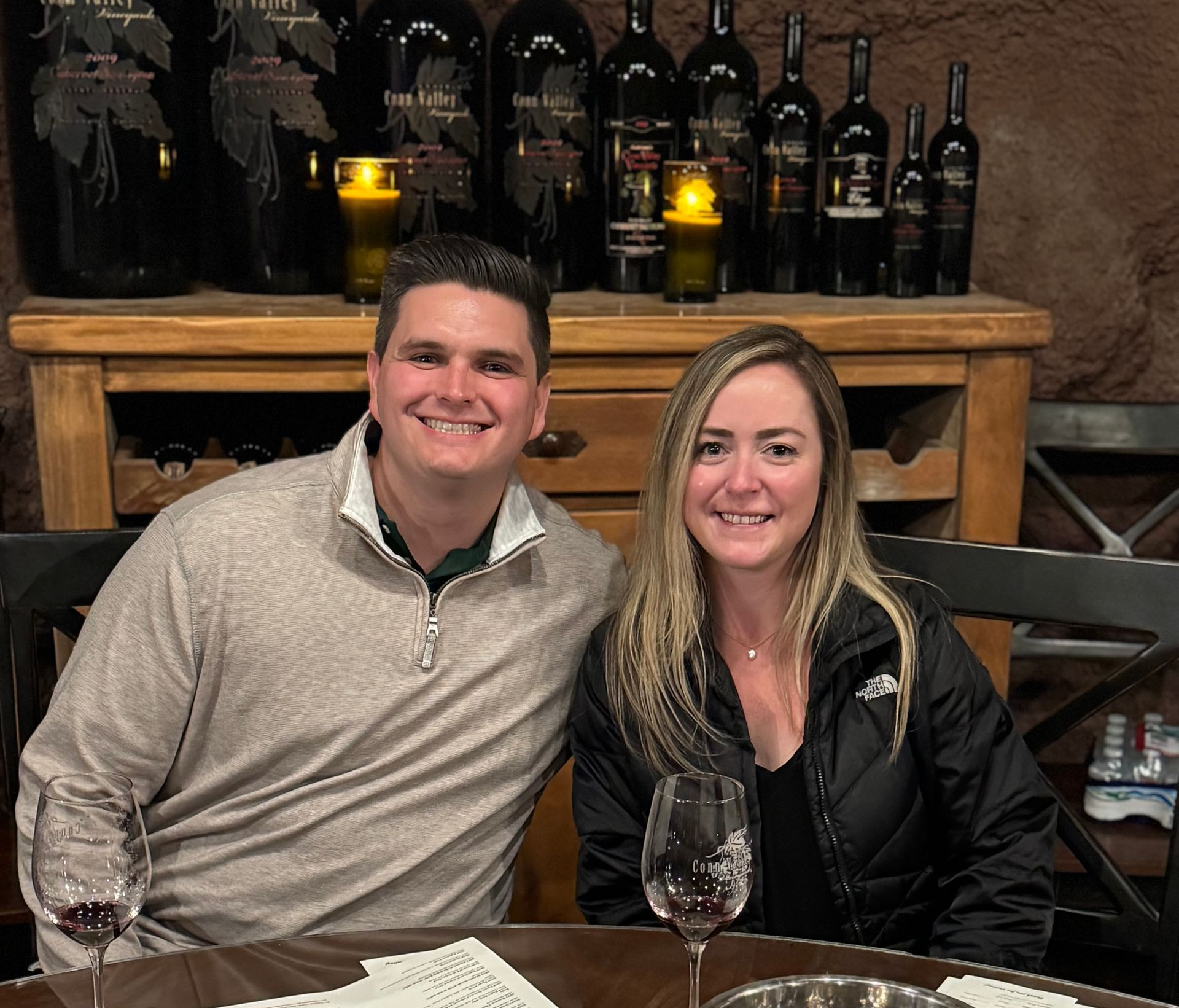 A couple sitting together at a table in a wine cellar with bottles on shelves in the background.
