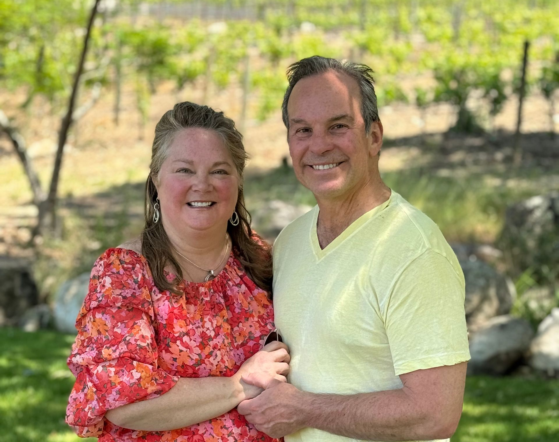 A smiling couple stands outdoors in front of a vineyard, holding hands.