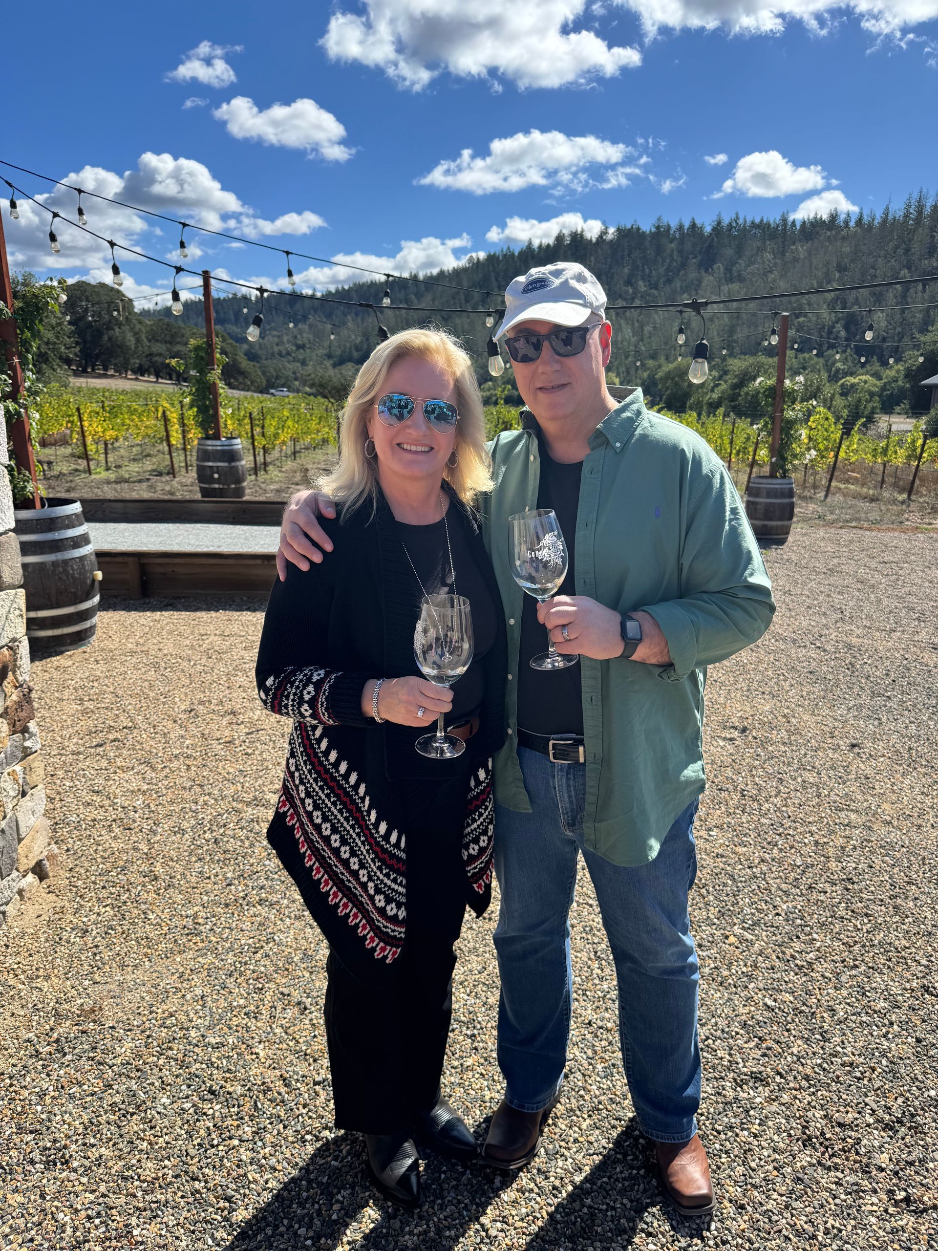 A smiling couple standing outdoors at a vineyard on a sunny day, each holding a wine glass.