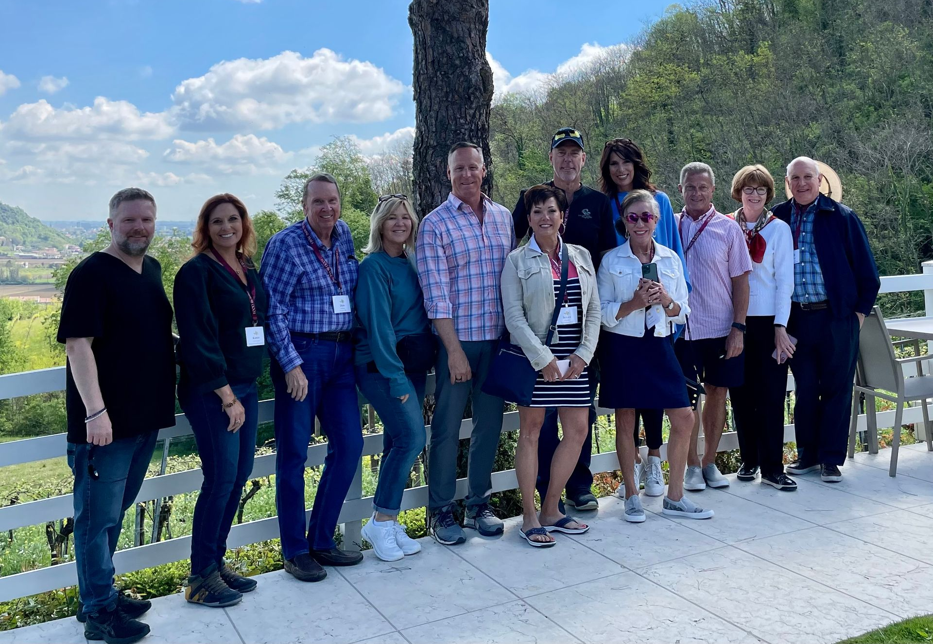 A group of thirteen people standing in a row outdoors on a sunny day at a vineyard with a white fence in the foreground.