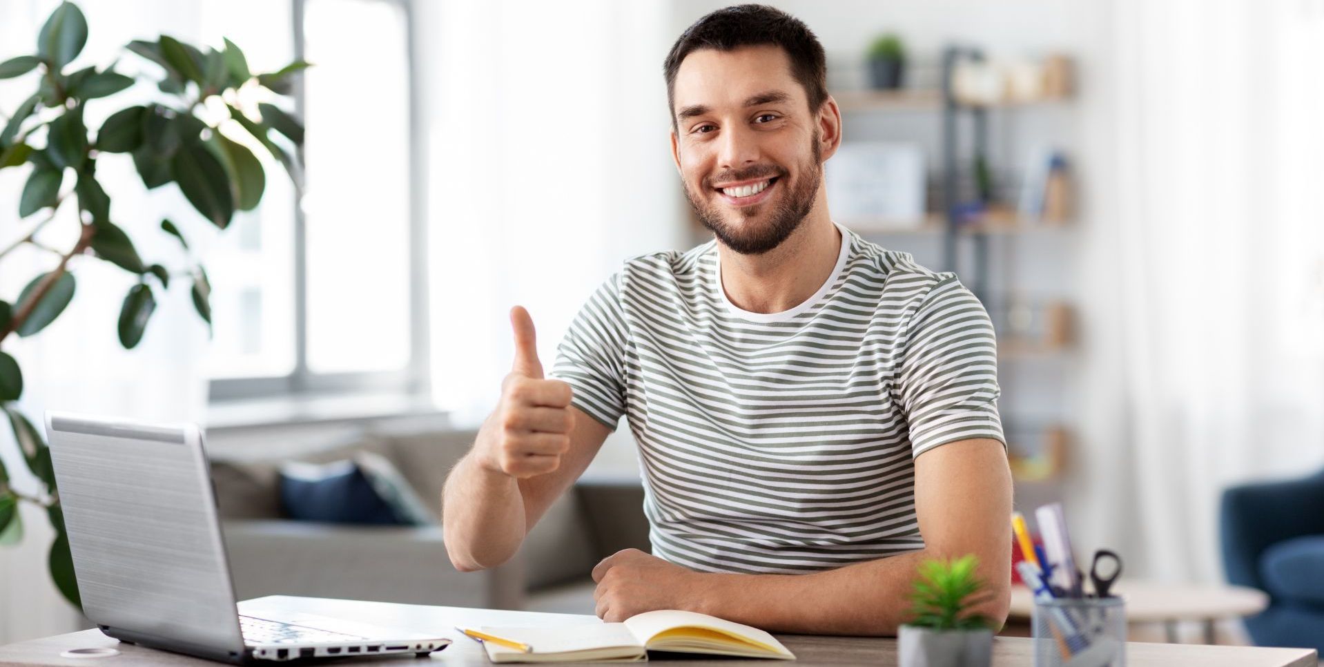 Man with thumbs up at a desk with a laptop, notepad, and potted plant.
