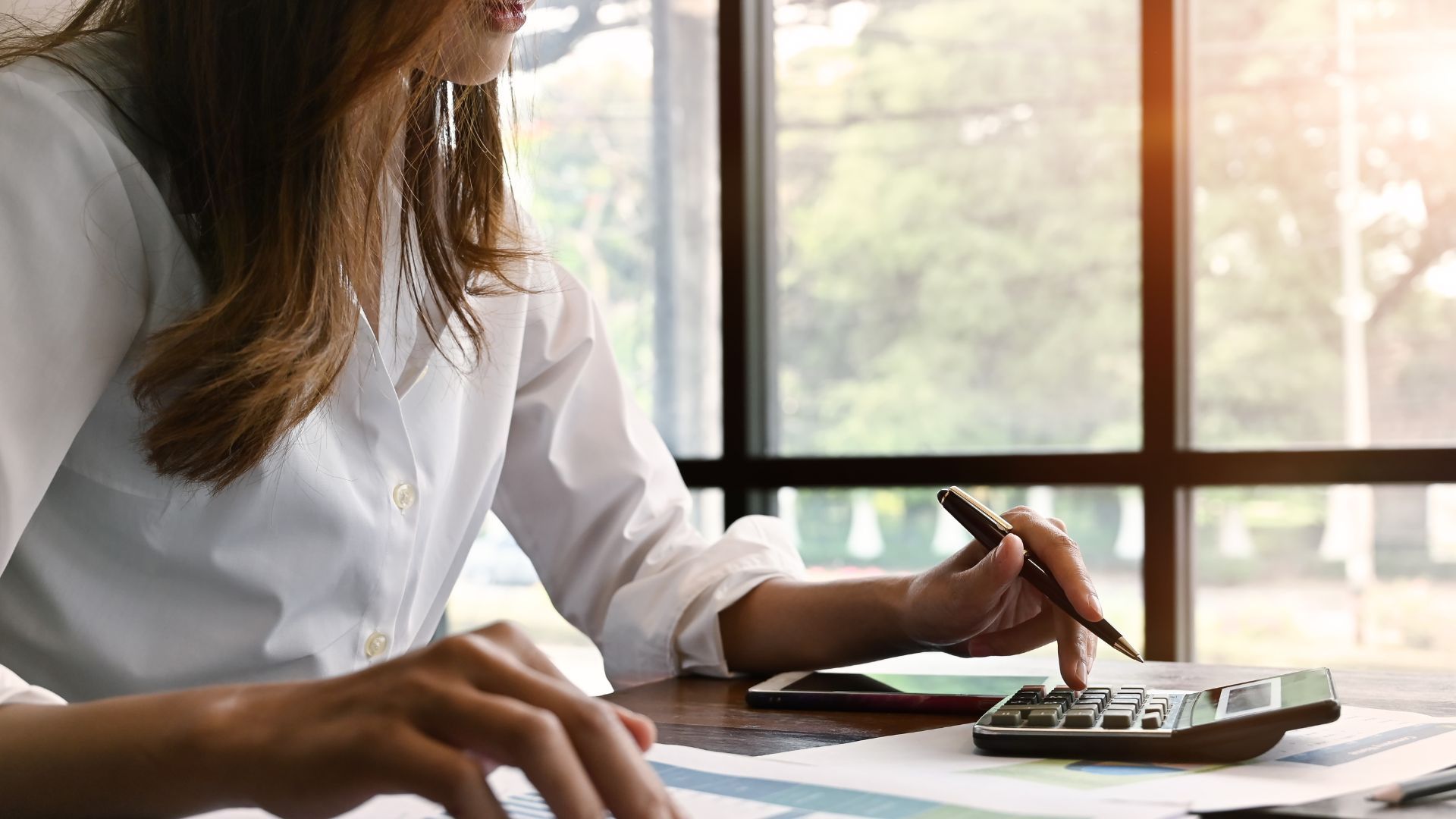 Woman in white shirt using calculator, at desk near window.