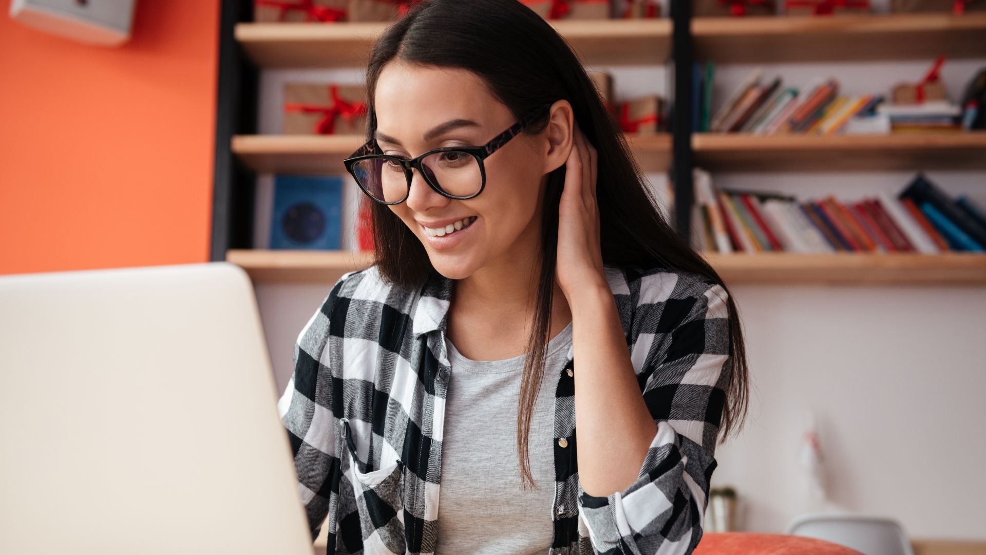 Woman with glasses, smiling at a laptop; wearing plaid shirt; bookshelves in the background.