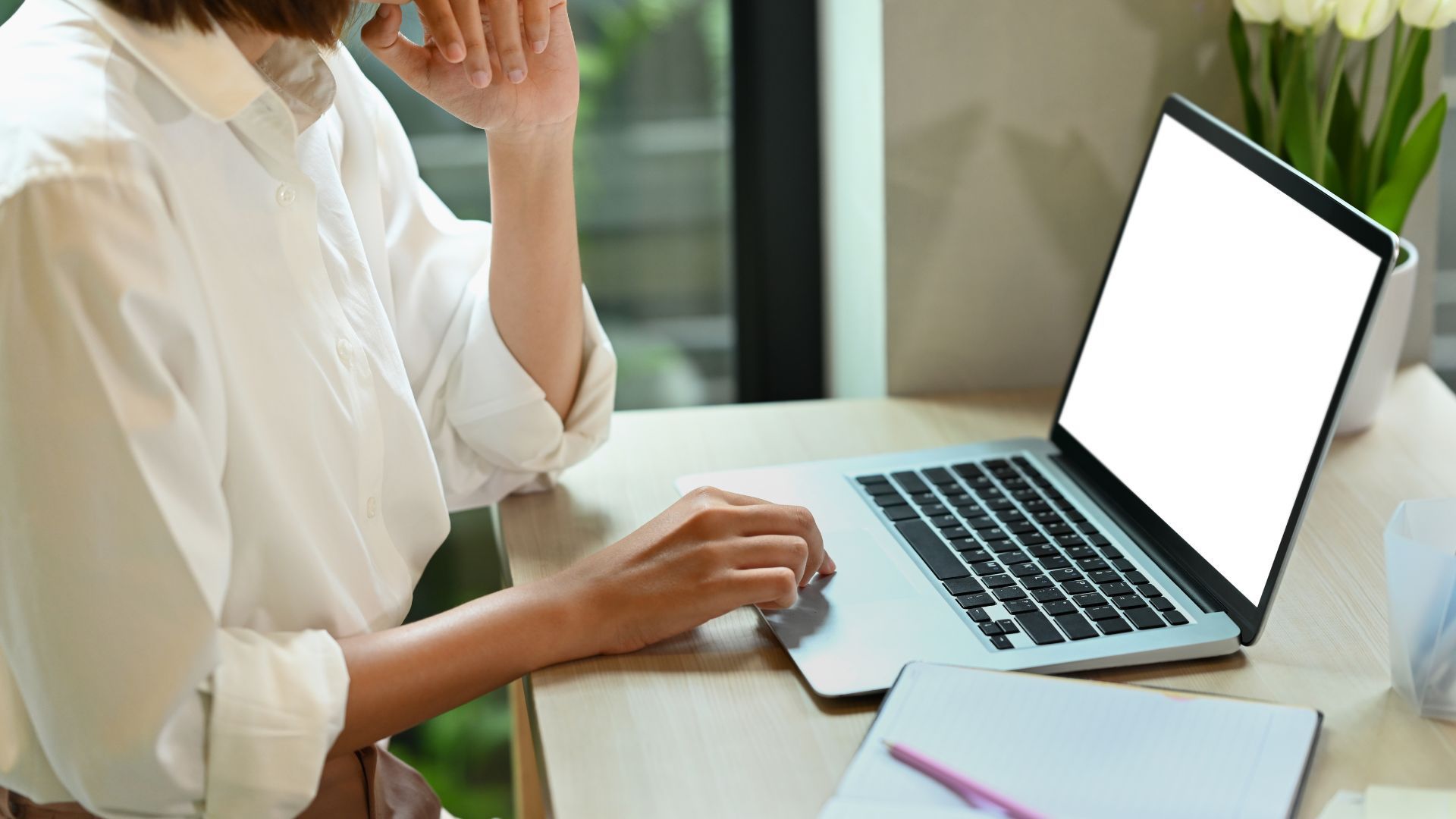Person in white shirt using laptop with blank screen at a desk with notepad and flowers.