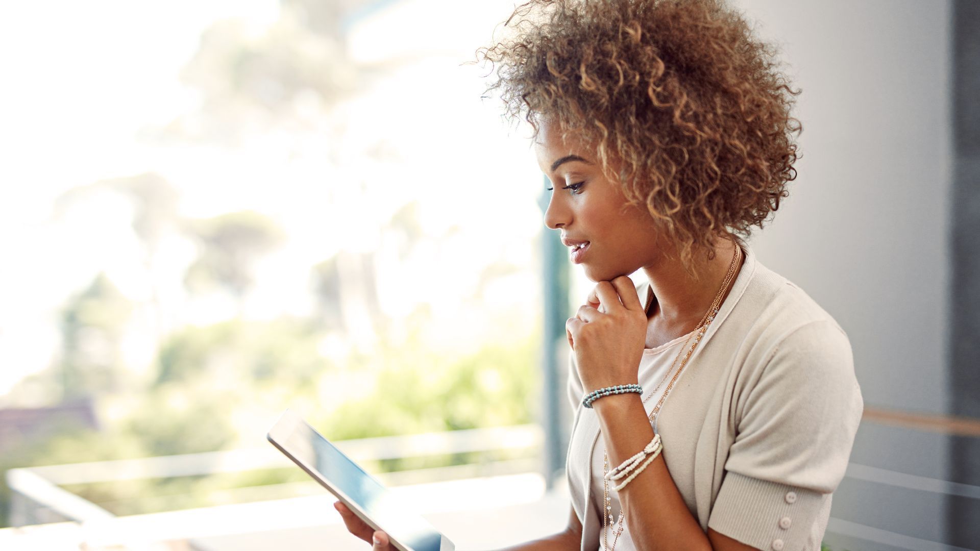 Woman with curly hair using a tablet, thoughtfully touching her chin near a window.