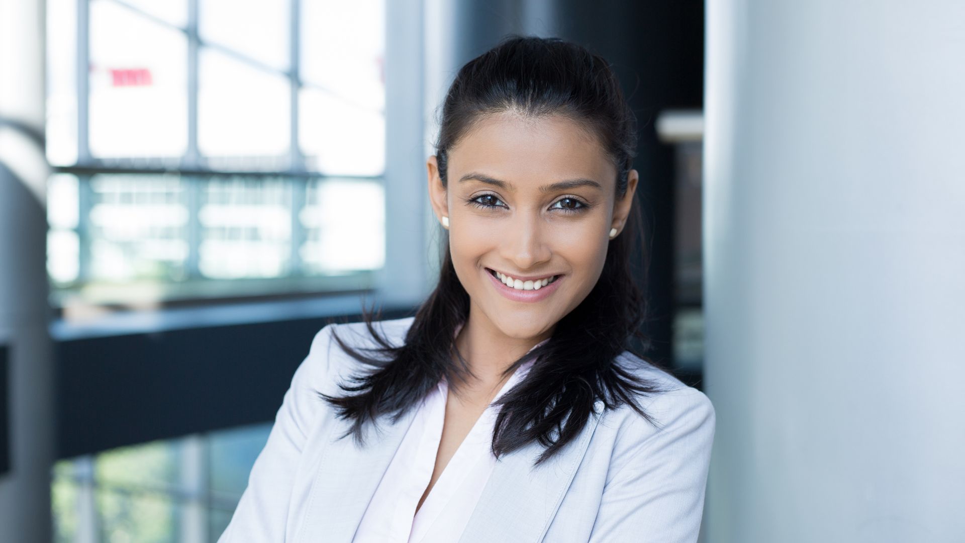 Woman in a white blazer smiles at the camera in a modern office setting.
