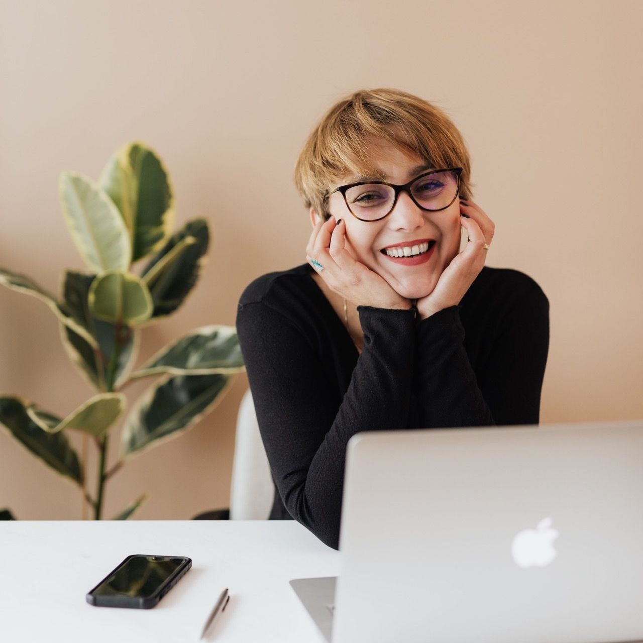 Woman with glasses smiling, hands on chin, in front of laptop and phone, next to plant in a bright room.