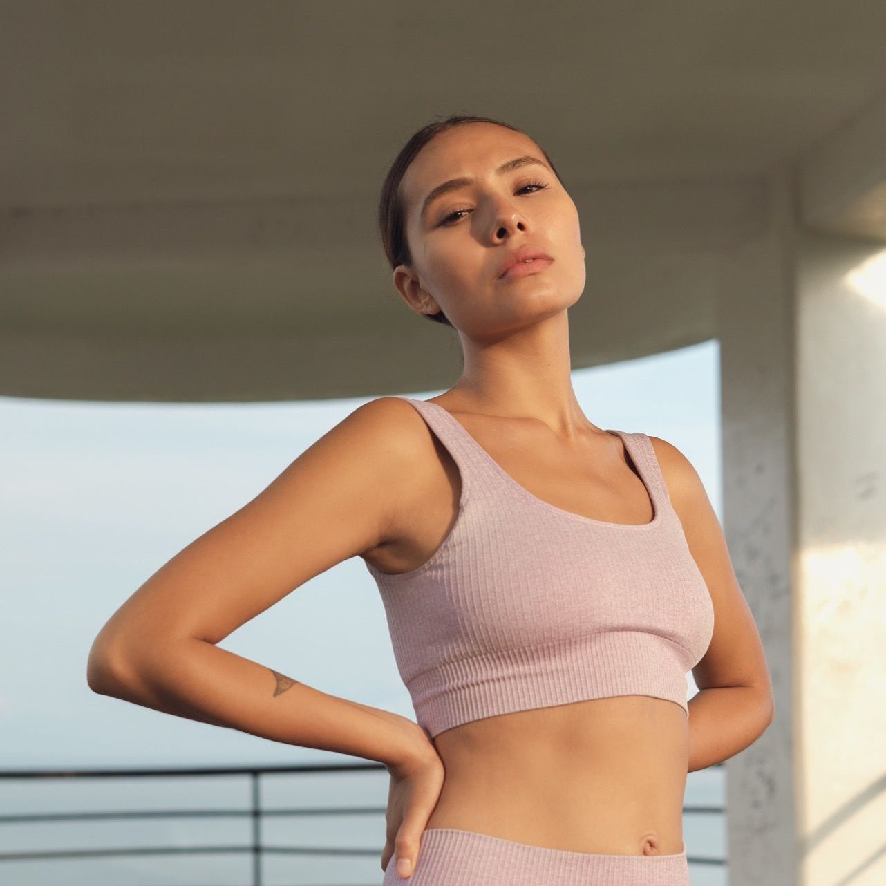 Woman in a pink sports bra poses outdoors with hands on hips, looking up.