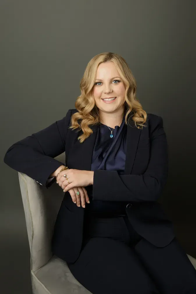 Smiling person in a black suit seated in a gray chair against a dark green background