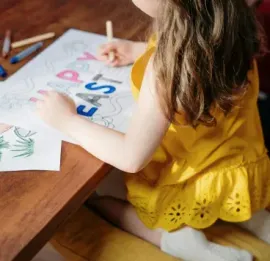 Child in a yellow dress drawing colorful letters at a wooden table.