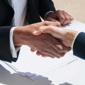 Two people in business suits shaking hands over a document on a desk