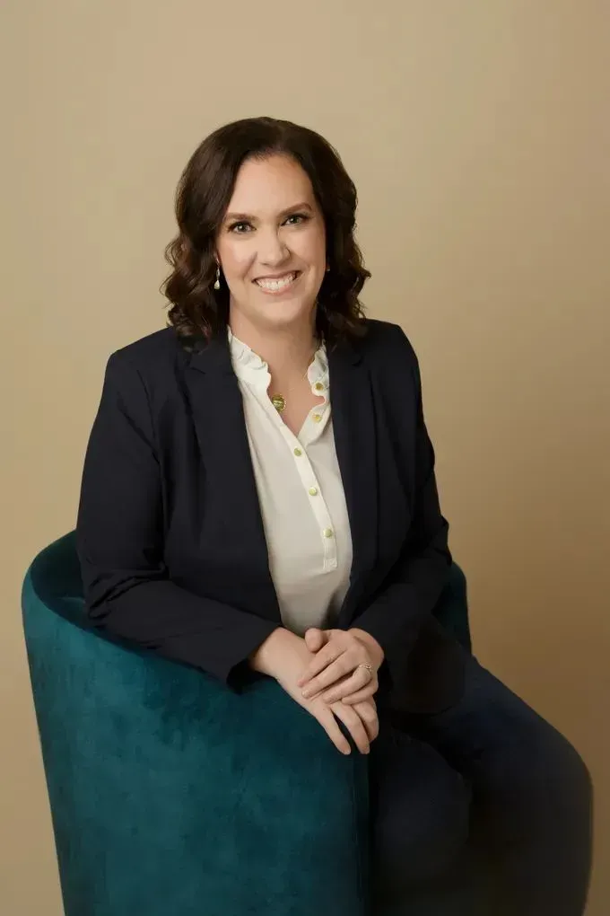 Woman seated in a teal chair, wearing a dark blazer and white blouse, smiling against a beige background