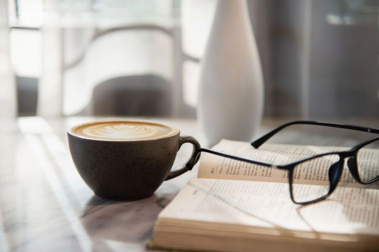 Black coffee cup beside glasses on an open book by a bright window