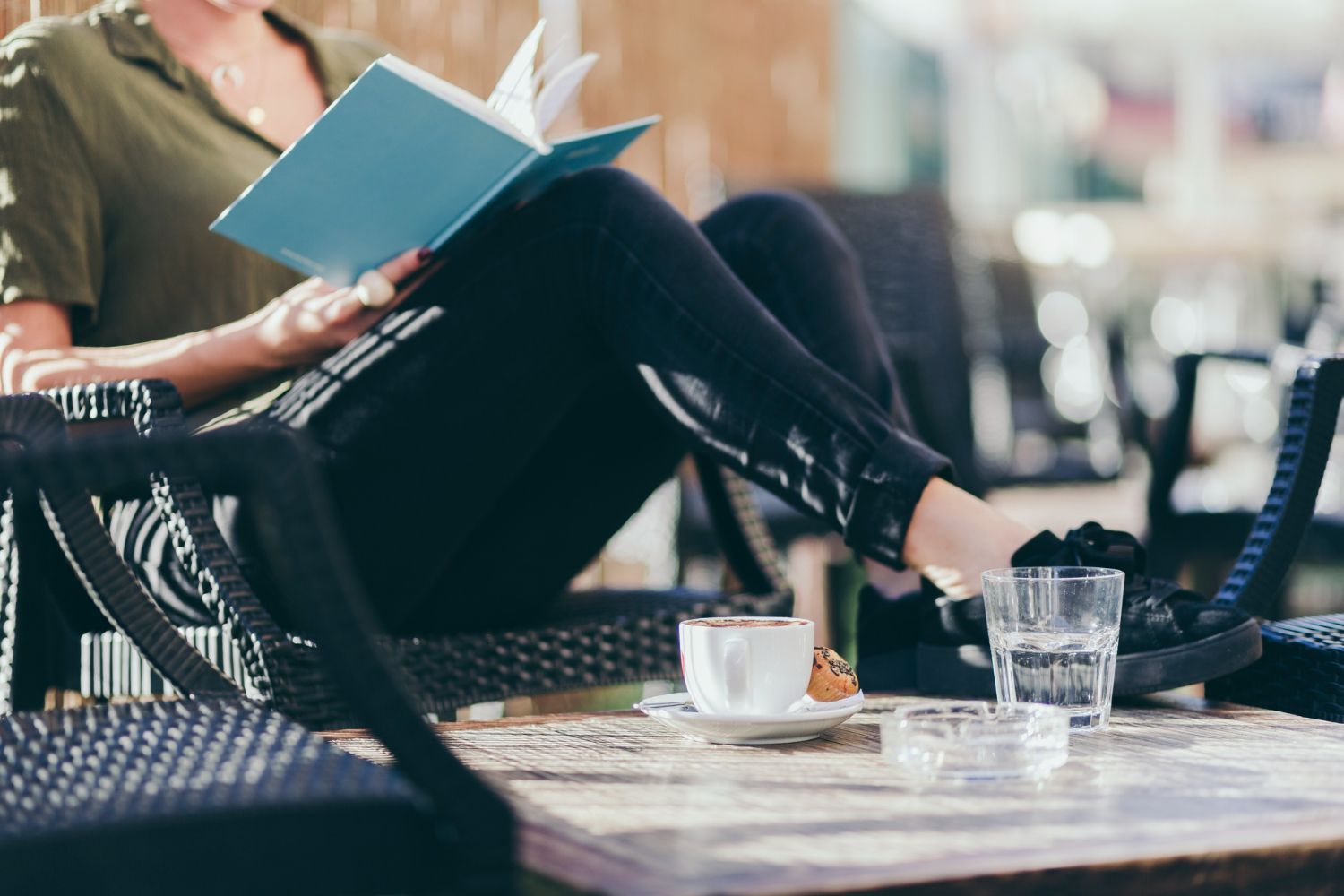 Person relaxing at a café, reading a blue book with a coffee cup and glass on the table.