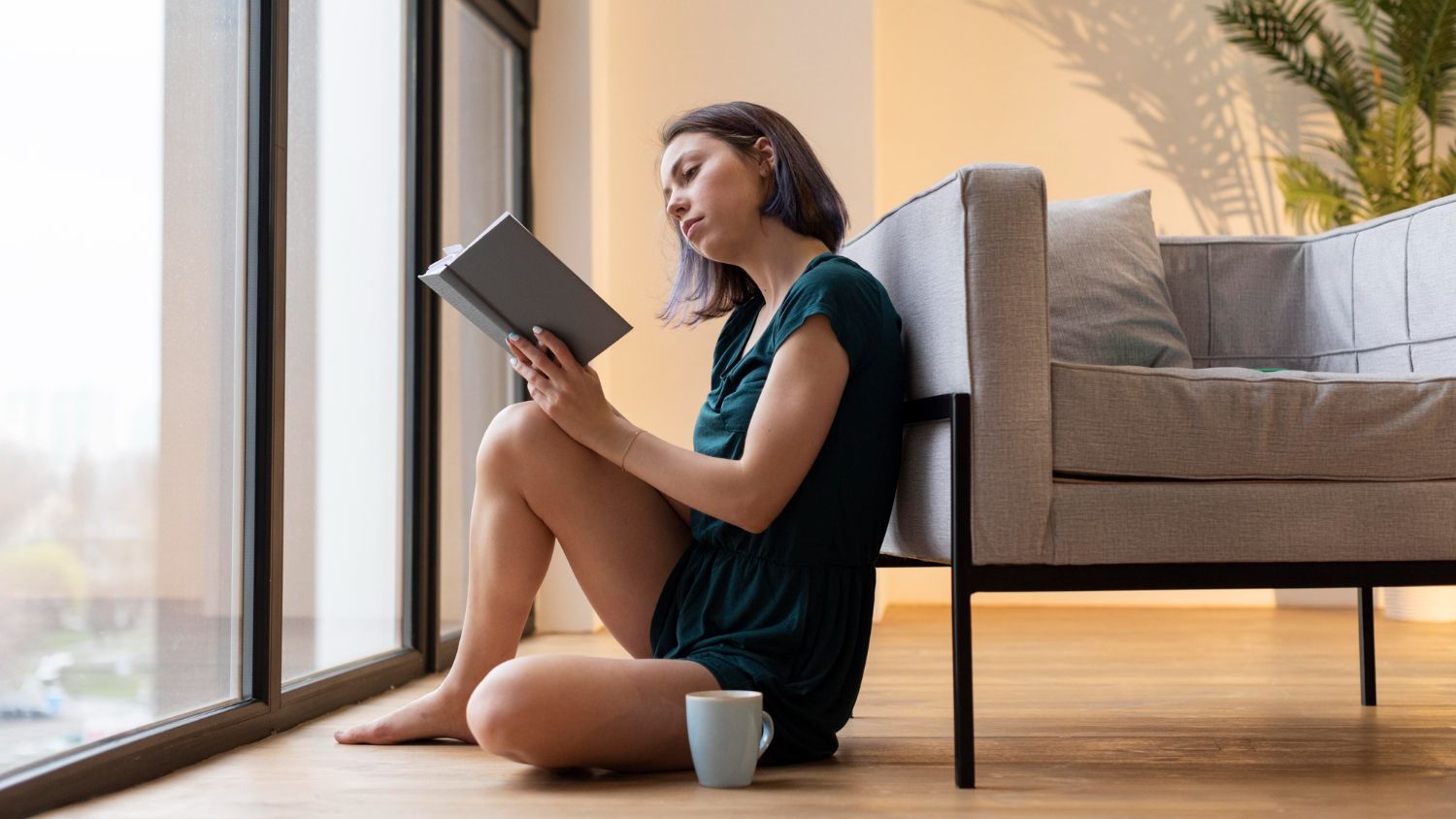 Person sitting by a window reading a book beside a sofa, with a coffee mug on the floor