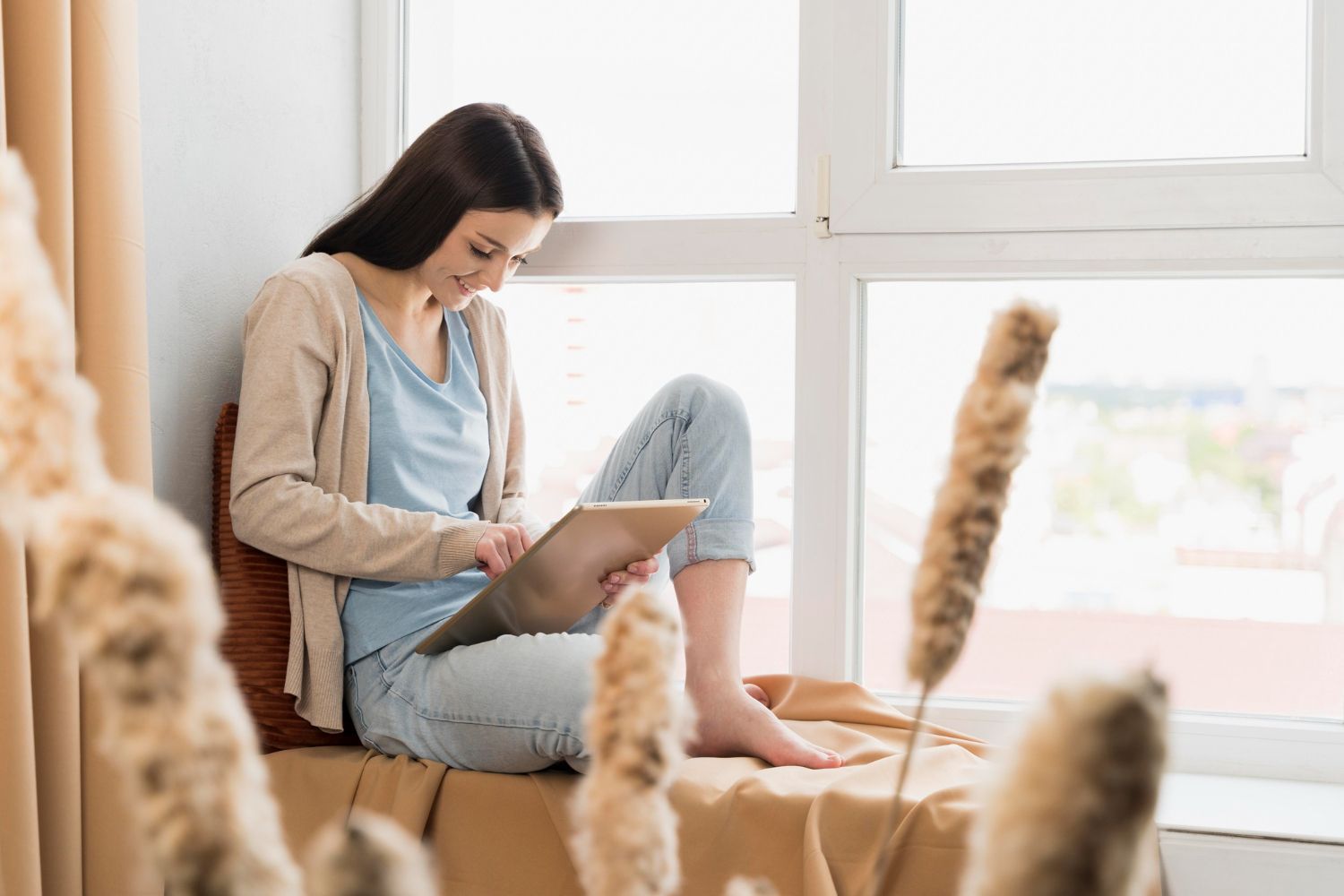 Woman sitting by a bright window, using a tablet on a couch with a cat in the foreground.
