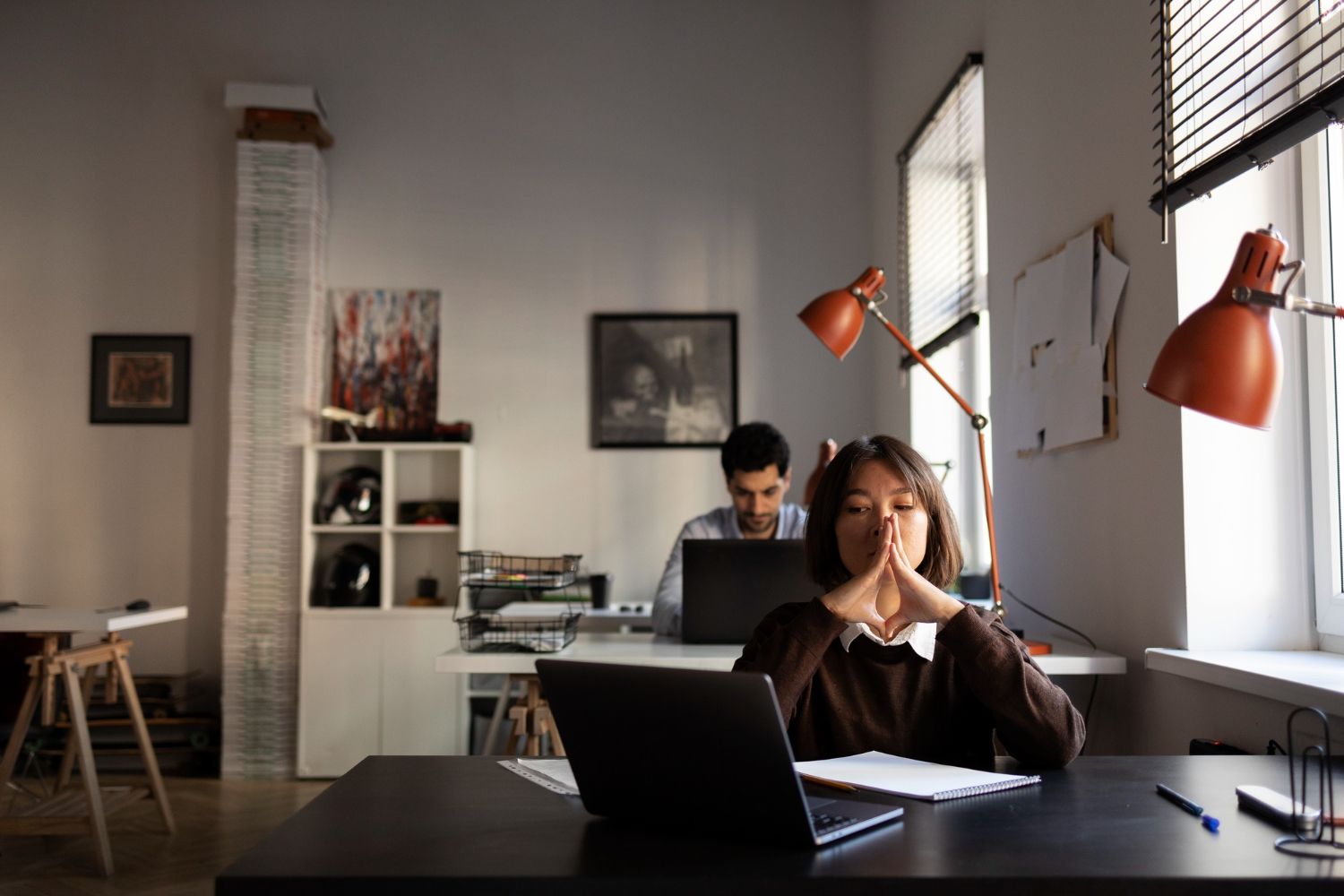 Two people working in a bright office, one in the foreground at a desk with a laptop and one in the back.